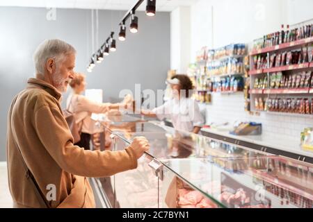 Junge lächelnde Verkäuferin mit lockigen Haaren, die der Dame Würstchen in einer Papierpackung aus dem Kühlschrank gab. Älterer Mann, der auf Glastheke mit großer Auswahl an Fleisch, Supermarkt. Stockfoto