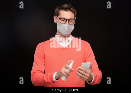 Junger Mann in Maske und Brille Desinfektion Smartphone mit Desinfektionsmittel vor schwarzem Hintergrund, Coronavirus-Konzept Stockfoto