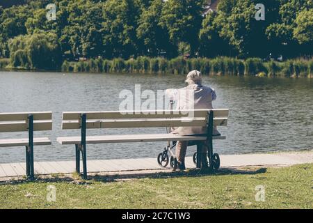 Ein älterer Mann sitzt mit seinem Spaziergänger auf einer Parkbank und schaut auf einen See Stockfoto