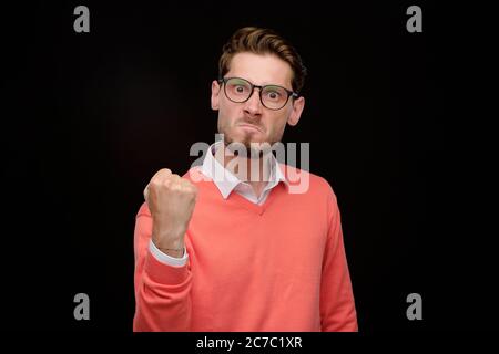 Porträt eines emotionalen jungen bärtigen Mannes, der Hand in der Faust hält und dabei seine Macht vor schwarzem Hintergrund zeigt Stockfoto