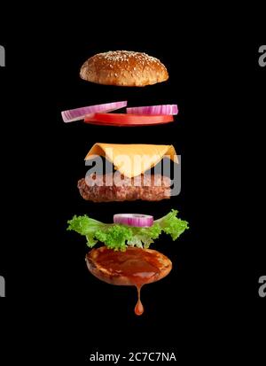 Fliegende Zutaten eines klassischen Käseeburger Sesambrötchen, Zwiebelringe, Tomatenscheiben und einem saftigen Grillschnitzel auf schwarzem Hintergrund. Fast Food Stockfoto