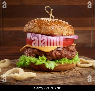 Cheeseburger mit Tomaten, Zwiebeln, Grillschnitzel und Sesambrötchen auf einem alten Holzschneidebrett, brauner Hintergrund. Fast Food Stockfoto