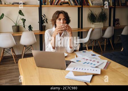 Junge schöne Frau sitzt am Tisch mit Laptop und Diagramme, während nachdenklich trinken Kaffee in modernen Büro Stockfoto