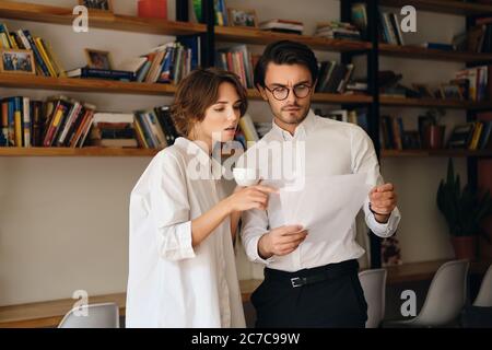 Zwei junge, nachdenkliche Kollegen stehen mit Papieren und diskutieren im Büro gemeinsam Stockfoto