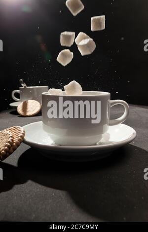 Anordnung von weißen Tassen, mehreren Keksen und schwebendem Zuckerwürfel. Stockfoto