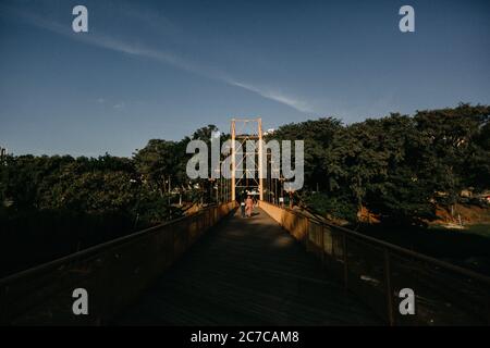 Horizontale Aufnahme einer Seilbrücke über einen Fluss in Ein Park mit zwei Personen, die darauf spazieren gehen Stockfoto
