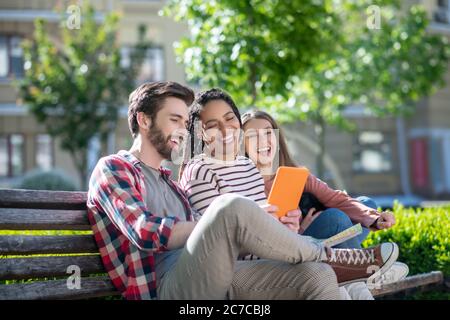 Guy und zwei Mädchen, die auf Tablet sitzen auf der Bank Stockfoto