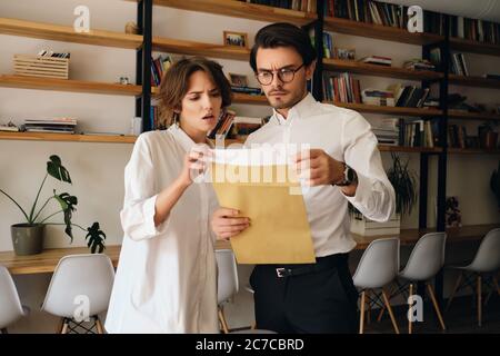 Junge enttäuschte Kollegen stehen nachdenklich beim Lesen Briefumschlag Brief während der Zusammenarbeit in modernen Büro Stockfoto