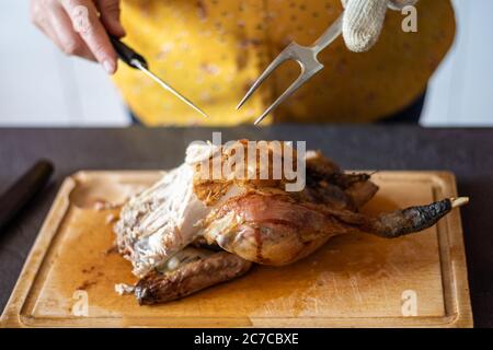 Kaukasische Frau, die ein gebratenes Huhn auf einer Holzplatte mit einem Messer und einer Gabel schnitzt. Stockfoto