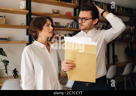Junge enttäuschte Kollegen aus dem Geschäft stehen mit Briefumschlag, während sie sich in modernen Büros traurig anguaschen Stockfoto