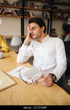 Junger müder Mann sitzt am Tisch mit Notizblock lehnte Kopf an der Hand schlafen bei der Arbeit in modernen Büro Stockfoto