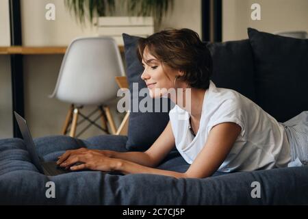 Seitenansicht der jungen attraktiven Frau auf dem Sofa liegend glücklich an neuem Projekt mit Laptop im Co-Working-Raum arbeiten Stockfoto