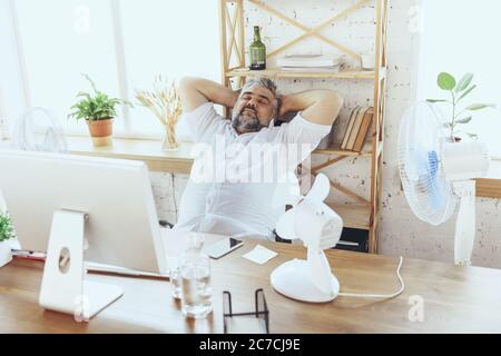 Müde. Geschäftsmann, Manager im Büro mit Computer und Ventilator, der sich abkühlt, heiß fühlt, gespült. Mit Ventilator, aber immer noch leiden von unbequem Klima im Schrank. Sommer, Büroarbeit, Business. Stockfoto