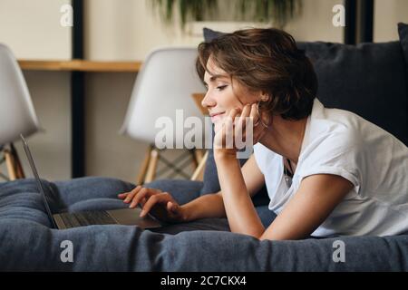 Seitenansicht einer jungen hübschen Frau, die auf dem Sofa liegt und auf dem Laptop im Co-Working-Bereich arbeitet Stockfoto