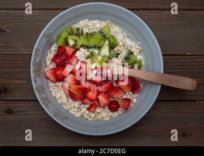 Haferflocken in einer braunen Platte mit gewürfelten Erdbeeren und Kiwi auf Holztisch, ganze Platte, zentriert, Draufsicht, mit Silberlöffel Stockfoto