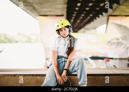 Teenager-Mädchen in Helm und stilvolle Kleidung posiert auf Half Pipe Rampe im Freien Skatepark. Schöne Kinder weibliche Modell Skateboarder mit Skate Board in Stockfoto