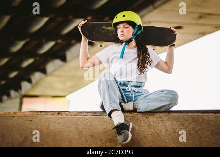 Teenager-Mädchen in Helm und stilvolle Kleidung posiert auf Half Pipe Rampe im Freien Skatepark. Schöne Kinder weibliche Modell Skateboarder mit Skate Board in Stockfoto
