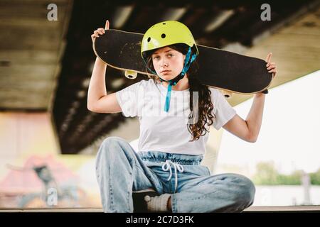 Teenager-Mädchen in Helm und stilvolle Kleidung posiert auf Half Pipe Rampe im Freien Skatepark. Schöne Kinder weibliche Modell Skateboarder mit Skate Board in Stockfoto