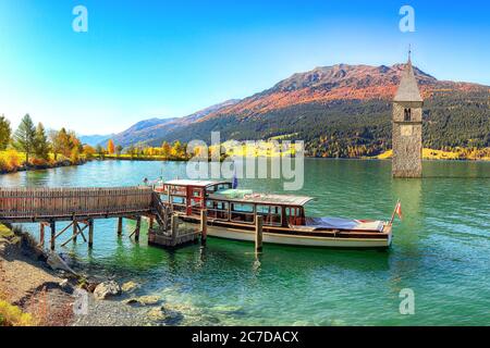 Fantastische Herbst Seenlandschaft mit einem Schiff in der Nähe der Pier und untergetauchten Glockenturm im See Reschen. Lage: Graun im Vinschgau Dorf, Reschensee Stockfoto