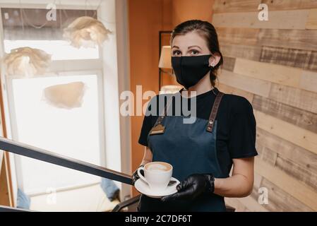 Eine Kellnerin, die eine schwarze Gesichtsmaske und Einweghandschuhe trägt, serviert in einem Restaurant eine Tasse Kaffee. Ein süßer Barista wartet auf Gäste in einer ca. Stockfoto