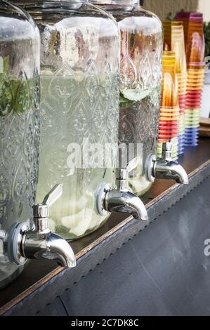 Vertikale Aufnahme von Glas Limonade Spender auf einem Holztisch Mit bunten Plastikbechern daneben Stockfoto