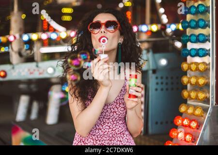Schöne Dame im Herzen Sonnenbrille und Kleid stehen und Blasen Blasen während der Zeit im Freizeitpark verbringen Stockfoto