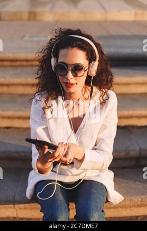 Schöne Dame mit dunklen lockigen Haaren in Sonnenbrille und weißer Jacke sitzt auf der Treppe mit Handy und Musik in Kopfhörer auf der Straße hören Stockfoto