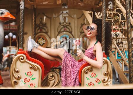 Fröhliche Mädchen mit dunklen lockigen Haaren in Sonnenbrille und Kleid hält Lully Pop Süßigkeiten in der Hand und Reiten auf Karussell, während glücklich Zeit in Stockfoto