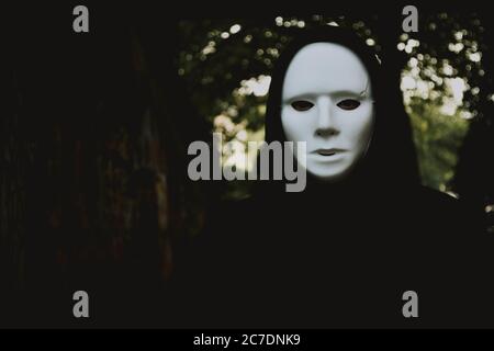 Selektive Nahaufnahme einer schwarzen Kapuzenperson in Weiß Maske auf dem verschwommenen Hintergrund von Bäumen Stockfoto
