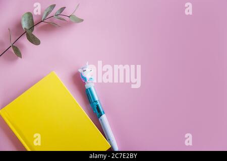 Gelbes Notizbuch, lustiger Einhorn-Stift und grüne Eukalyptusblätter auf rosa Pastellhintergrund. Flach liegend. Draufsicht. Speicherplatz kopieren Stockfoto