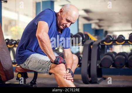 Ein Porträt von kahlem älteren Mann, der während des Trainings im Fitnessstudio starke Knieschmerzen empfinden kann. Menschen, Gesundheit und Lifestyle-Konzept Stockfoto