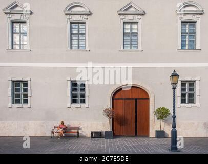 Budapest, Ungarn, August 2019, Frau sitzt auf einer Bank und liest vor einem Gebäude im Burgviertel Stockfoto