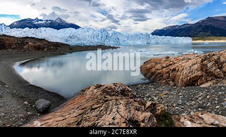 Perito Moreno Gletscher, der sich im Gletschersee vor ihm spiegelt, mit Felsen im Vordergrund und Bergen im Hintergrund Stockfoto