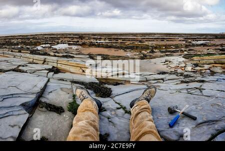 Herrenbeine und Stiefel mit Hammer und Meißel ausgestreckt an der Seite an einer britischen Schieferküste bei Ebbe mit Ammoniten Fossilien im Felsen platziert Stockfoto