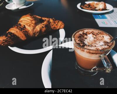 Nahaufnahme einer Tasse aus klarem Glas, gefüllt mit Cappuccino Neben einem Croissant Stockfoto