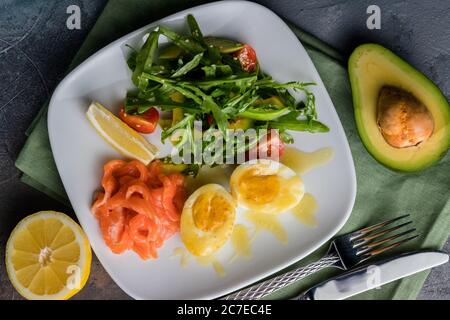 Salat mit gesalzener Forelle, Avocado, Rucola und Spiegelei in Butter. Besteck auf grauem Hintergrund Stockfoto