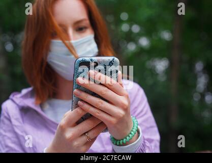 Redhead Mädchen in Medizin Maske hält ein Smartphone und Nachrichten lesen Stockfoto
