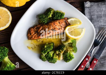 Gegrilltes Forellensteak auf einem weißen Teller mit Brokkoli und Limette. Besteck auf dem Holzhintergrund Stockfoto