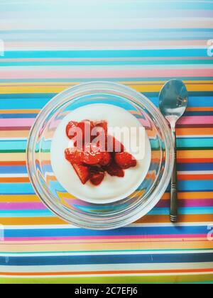 Joghurt in einer Glasschüssel mit Erdbeeren oben daneben Ein silberner Löffel auf einer bunt gestreiften Oberfläche Stockfoto