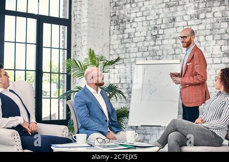 Selbstbewusster Geschäftsmann, der auf Flipcharts schreibt, präsentiert das neue Projekt in der Vorstandsetage bei der Unternehmenssitzung. Serious Mentor Auditor spricht mit Partnern Stockfoto