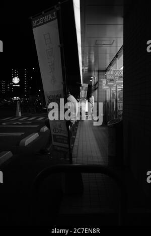 MATSUYAMA, JAPAN - 23. Sep 2019: Eine vertikale Graustufenaufnahme von Menschen auf der Straße, die nachts auf den Bus warten Stockfoto