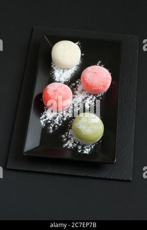 Vier Arten von japanischen Dessert-Mochi - Granatapfel mit Honig, grüner Matcha-Tee, Erdbeere, Kokosnuss auf einem schwarzen Teller auf einem schwarzen Tisch. Nahaufnahme Stockfoto