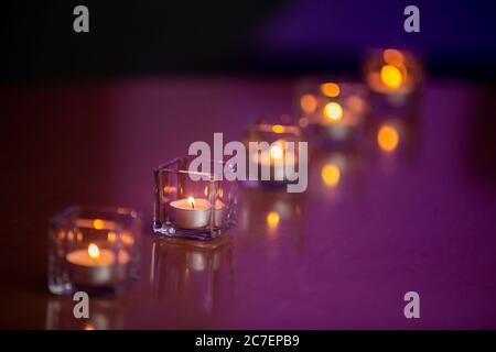 Entzündete Teelichtkerzen in Glasbehältern auf einer glänzenden Tischplatte mit einem violetten Hintergrund Stockfoto