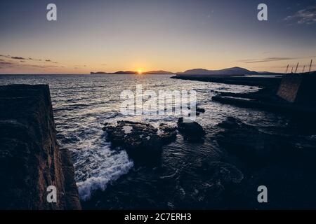 High-Angle-Aufnahme von Meereswellen, die auf die Felsen treffen Das Ufer mit einer Sonne, die hinter den Bergen scheint Stockfoto