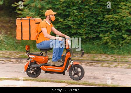 Ein Mann mit Lebensmittelzustellung in Orange mit einer schützenden medizinischen Maske auf seinem Gesicht reitet ein Moped mit einem Lebensmittelzulieferbeutel. Lebensmittelzustellung während der Isolierung Stockfoto