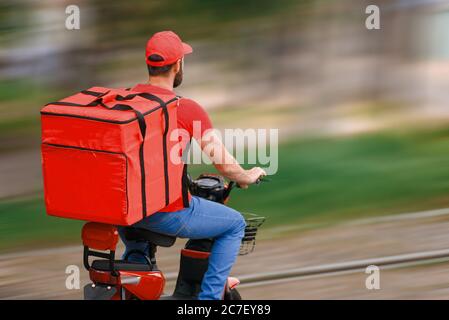 Ein Lebensmittellieferer in roter Uniform fährt mit einem Moped und einer Lebensmittelverpackung. Blick von hinten. Stockfoto