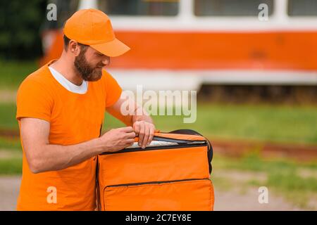 Der Lebensmittellieferer in orangefarbener Uniform öffnet einen Lebensmittellieferbeutel. Stockfoto