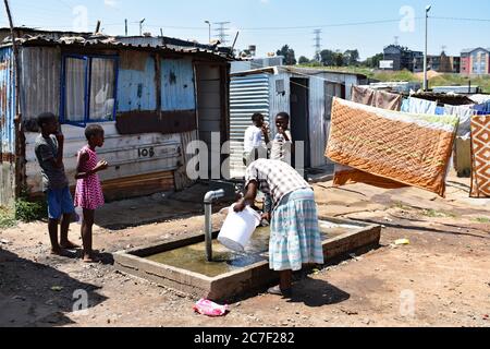 Eine Mutter füllt einen Eimer Wasser aus einem Wasserhahn, während ihre Kinder in einem Slum in Soweto, Johannesburg, auf sie schauen. Kleidung und Bettwäsche trocknen auf der Wäscheleine Stockfoto