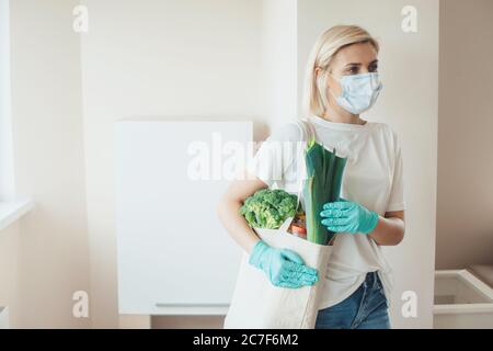 Blonde kaukasische Frau trägt eine medizinische Maske auf Gesicht und Handschuhe hält eine Tasche mit Produkten Stockfoto