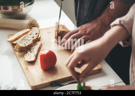 Kaukasischer Mann, der Brot aufschneiden muss, während seine Frau in der Küche einen Salat aus Gemüse zubereitet Stockfoto
